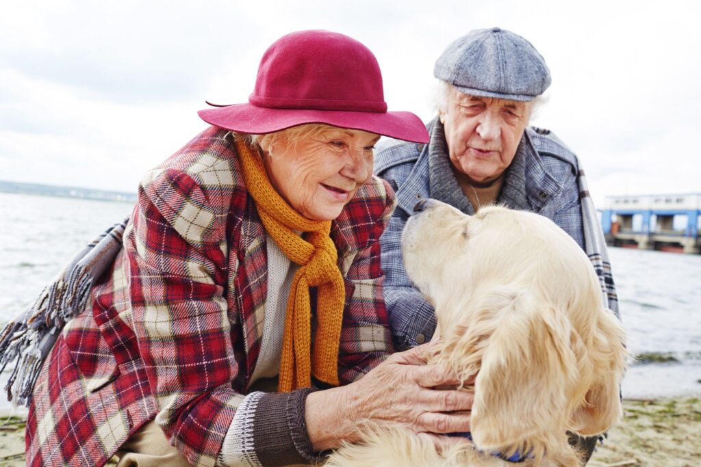 Elderly man and woman petting a Golden Retriever outdoors near the water, illustrating companionship, emotional support, and physical engagement for older adults