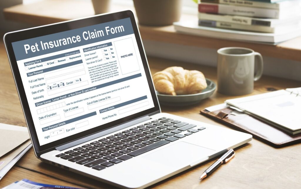 Open laptop on a wooden desk displaying a pet insurance claim form, with a coffee mug, croissant on a plate, books, pen, and notepad in the background, bright home office setting.