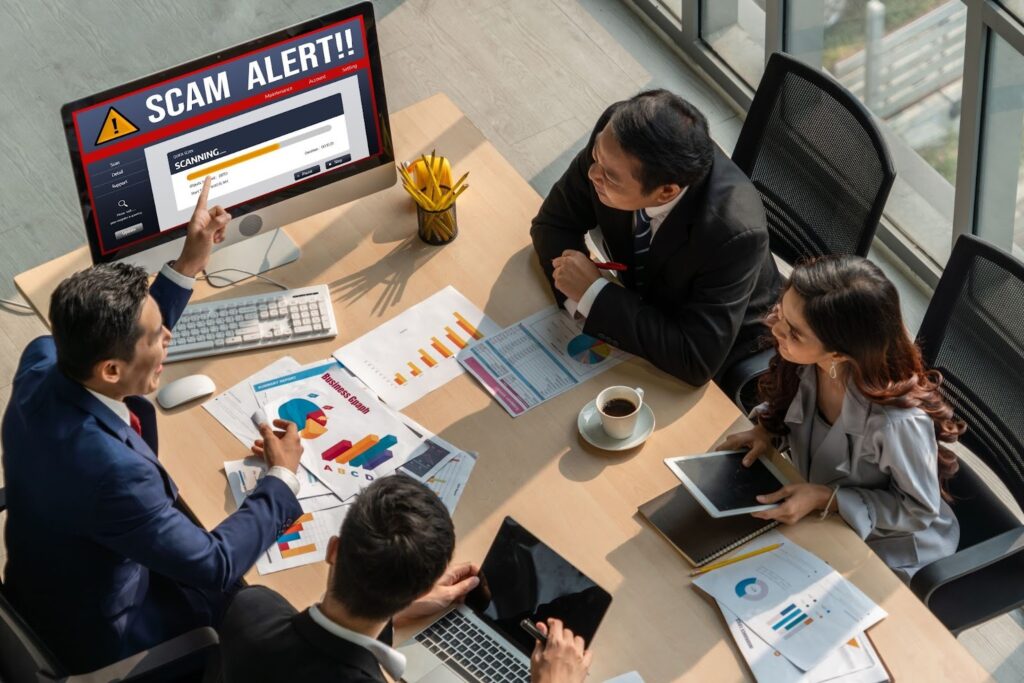 Four business people sitting around a table in a meeting room, reviewing charts and documents, while a computer screen in front displays a 'SCAM ALERT!!' warning message.