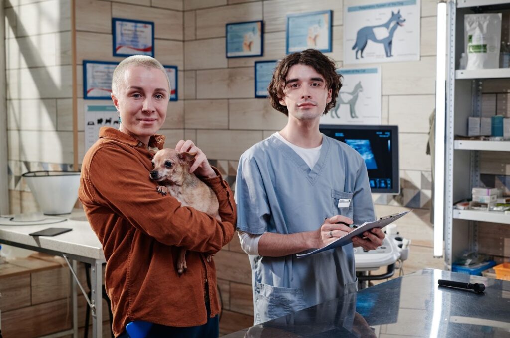 Veterinarian holding a clipboard and a pet owner holding a small Chihuahua in a veterinary clinic, with medical charts and supplies visible in the background.