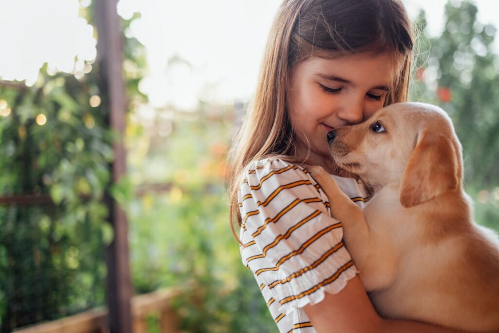 Young girl holding and cuddling a golden Labrador puppy outdoors, smiling as the puppy nuzzles her face.